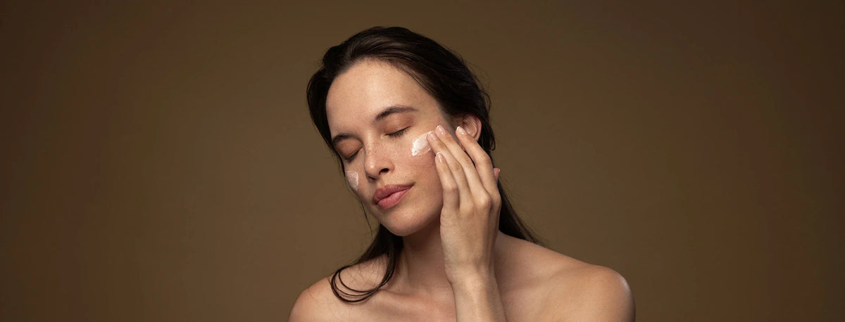 Woman applying Antipodes moisturiser to her cheek against a brown gradient background.