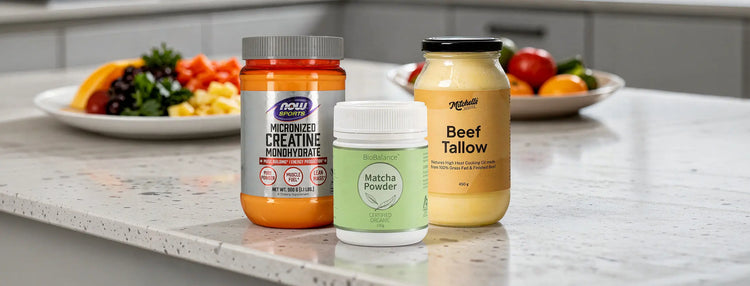 Three supplement containers arranged on a kitchen countertop: a large orange jar labelled Micronized Creatine Monohydrate, a small white jar labeled Matcha Powder, and a yellow jar labeled Beef Tallow, with blurred fruit and vegetables in the background.