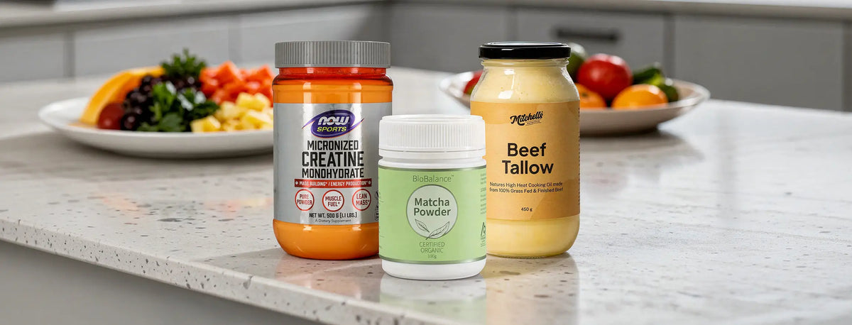 Three supplement containers arranged on a kitchen countertop: a large orange jar labelled Micronized Creatine Monohydrate, a small white jar labeled Matcha Powder, and a yellow jar labeled Beef Tallow, with blurred fruit and vegetables in the background.