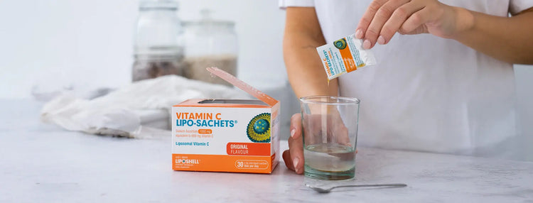 Person preparing a glass of water with vitamin C lipo-sachets on a white surface.