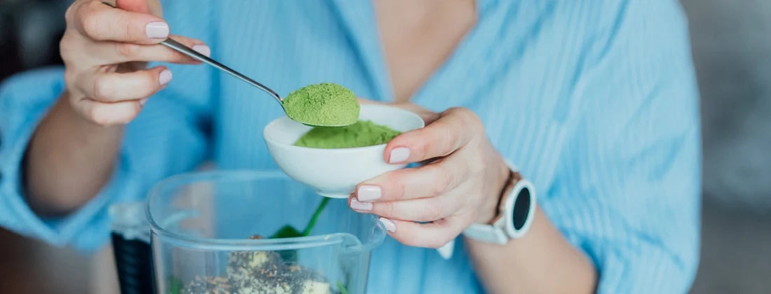Woman wearing a blue shirt and holding a small bowl with green powder and spooning some into a smoothie blender.