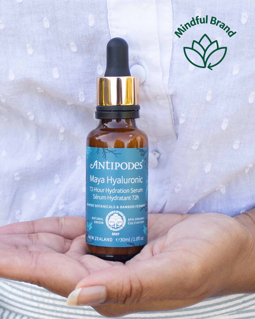 Antipodes Maya Hyaluronic serum bottle held in hands with 'Mindful Brand' logo in the corner.