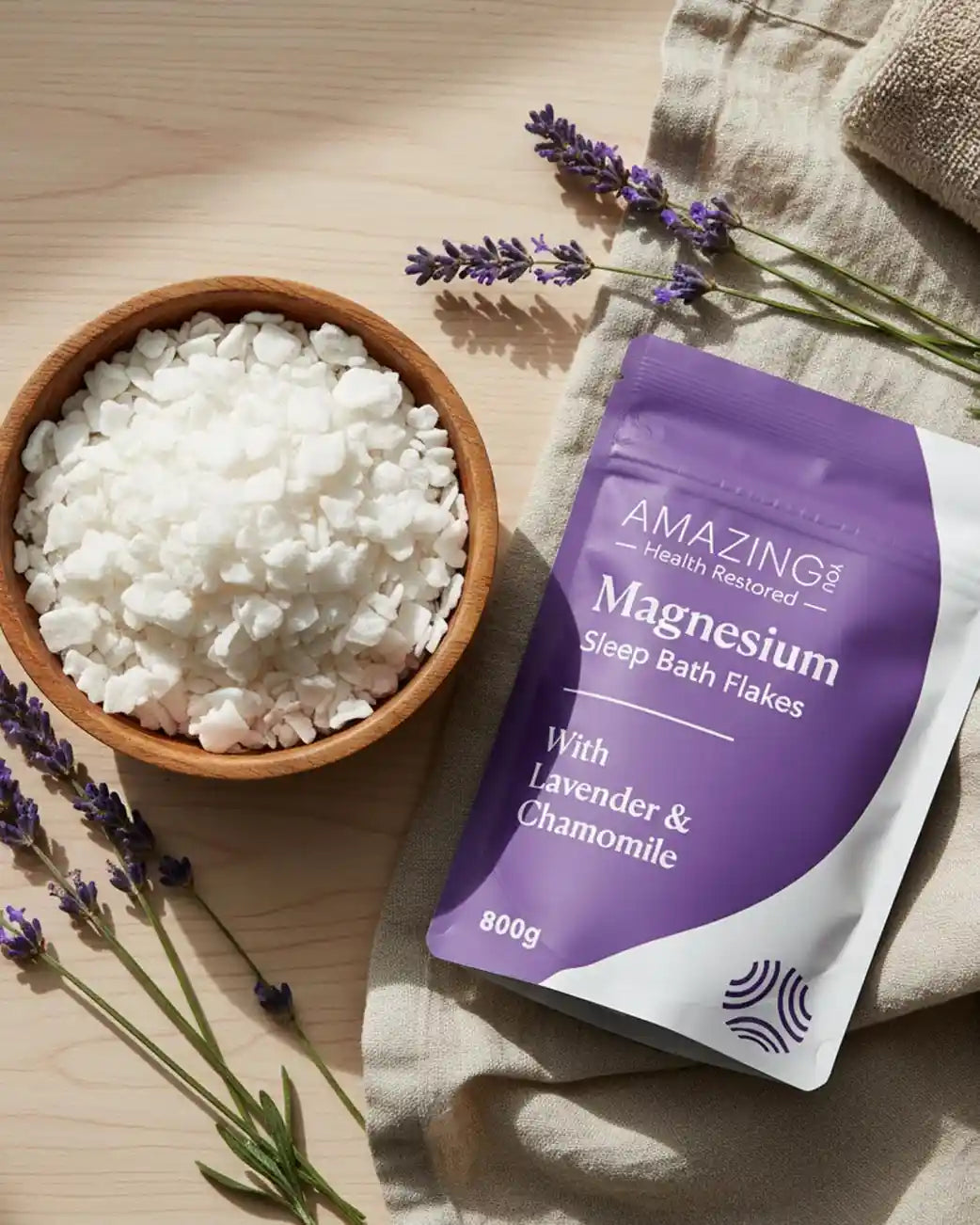 Bowl of magnesium bath flakes with a pouch of Amazing You Magnesium Sleep Bath Flakes on a wooden surface with lavender.
