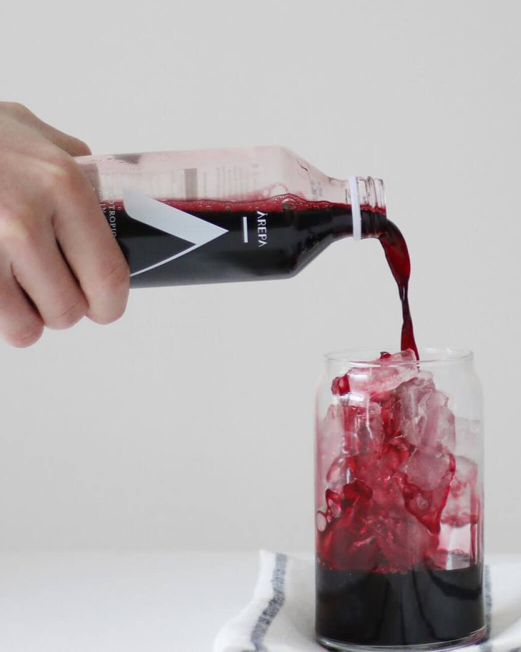 Hand pouring a dark red Arepa beverage from a bottle into a glass with ice on a white background