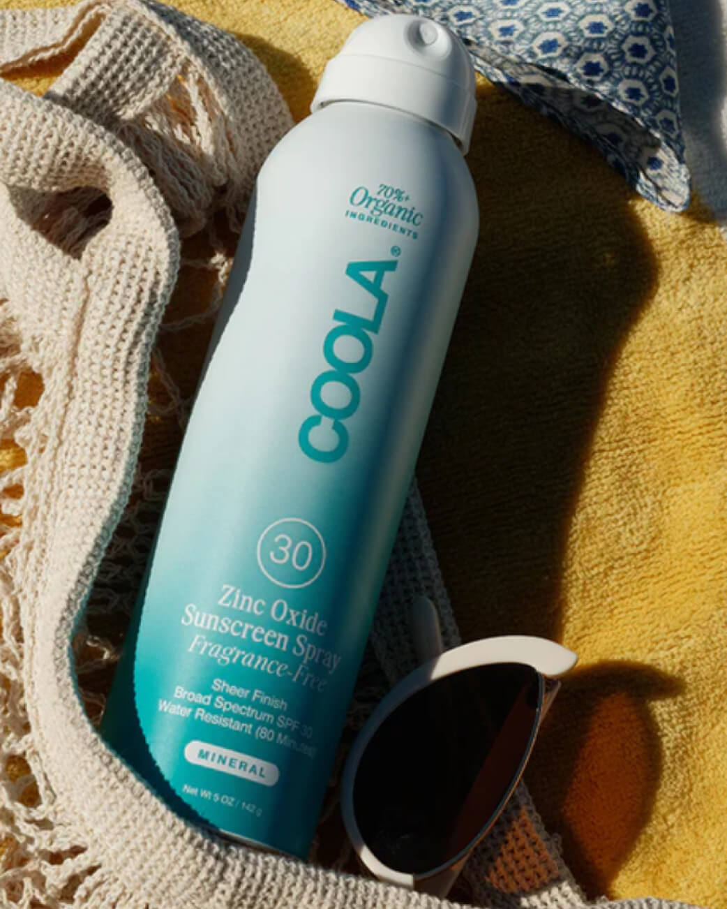 Coola sunscreen spray bottle on a textured surface with sunglasses and a towel.