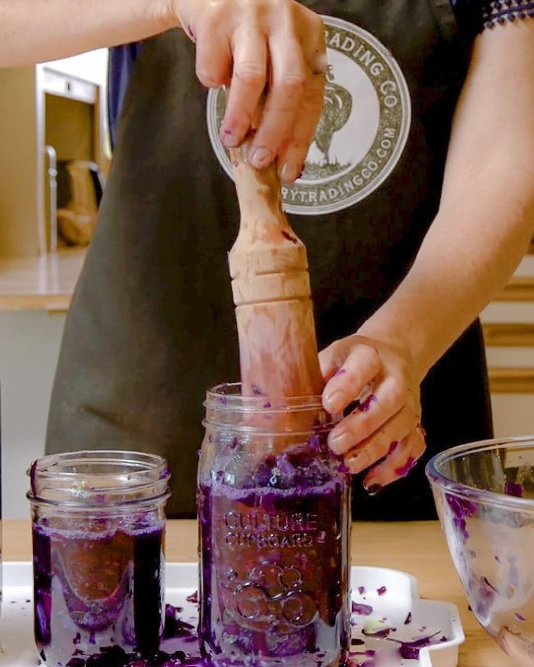 Person using a wooden spoon to stir purple liquid in a mason jar, with a visible brand logo on their apron.