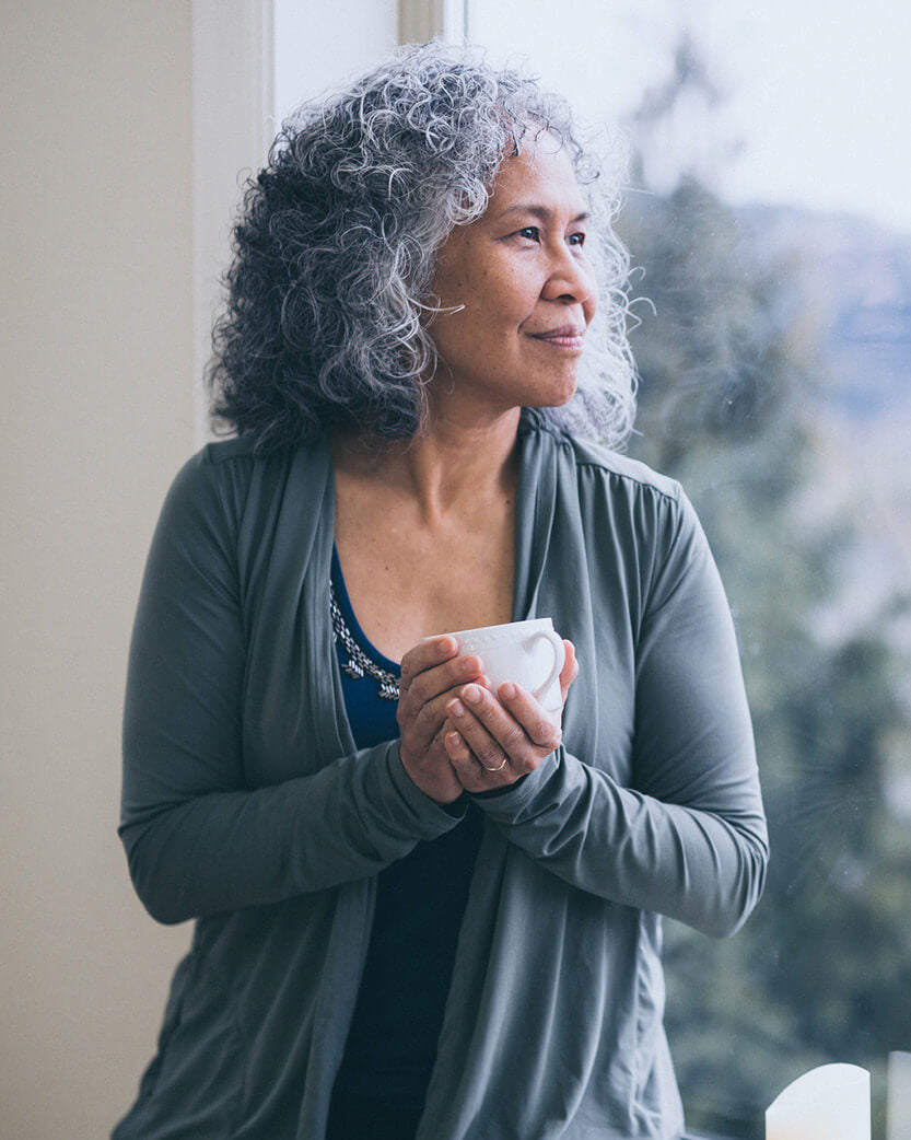 Woman holding a mug, looking out a window with a blurred outdoor background.