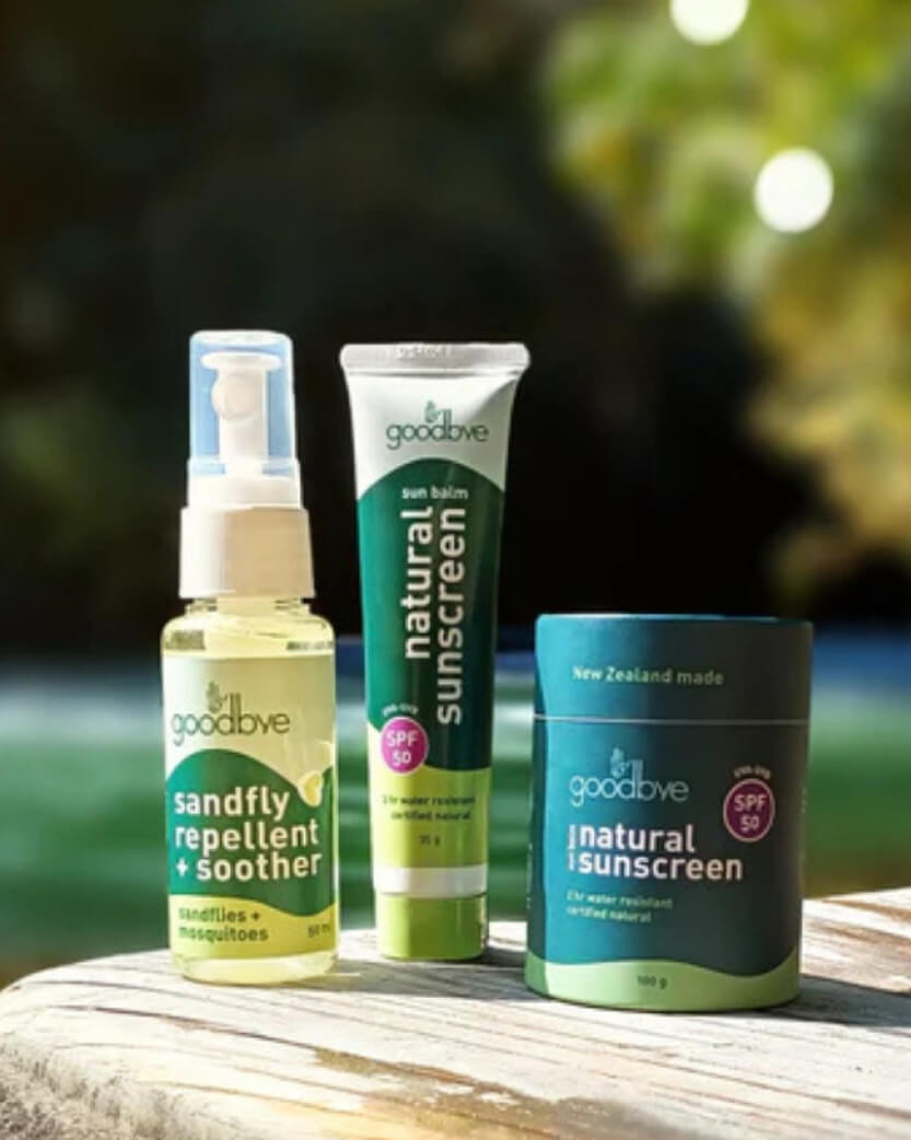 Three Goodbye products including a spray bottle, tube, and container on a wooden surface with a blurred natural background.