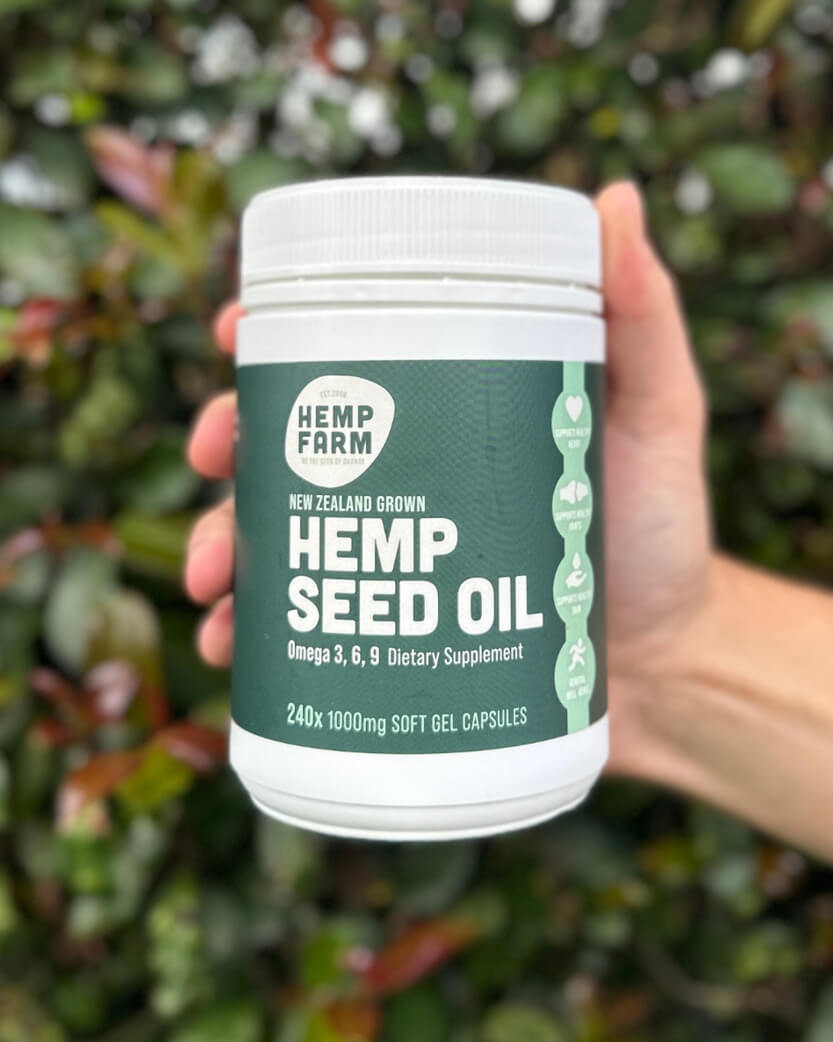 Hand holding a container of Hemp Farm hemp seed oil capsules with a green leafy background.