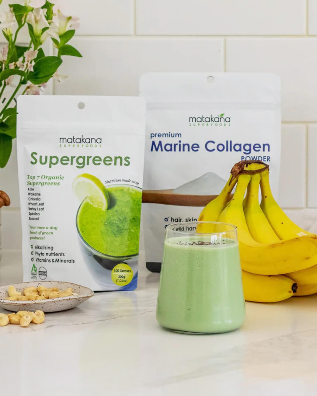 Supergreens and Marine Collagen powder packages with a green smoothie and bananas on a kitchen counter.