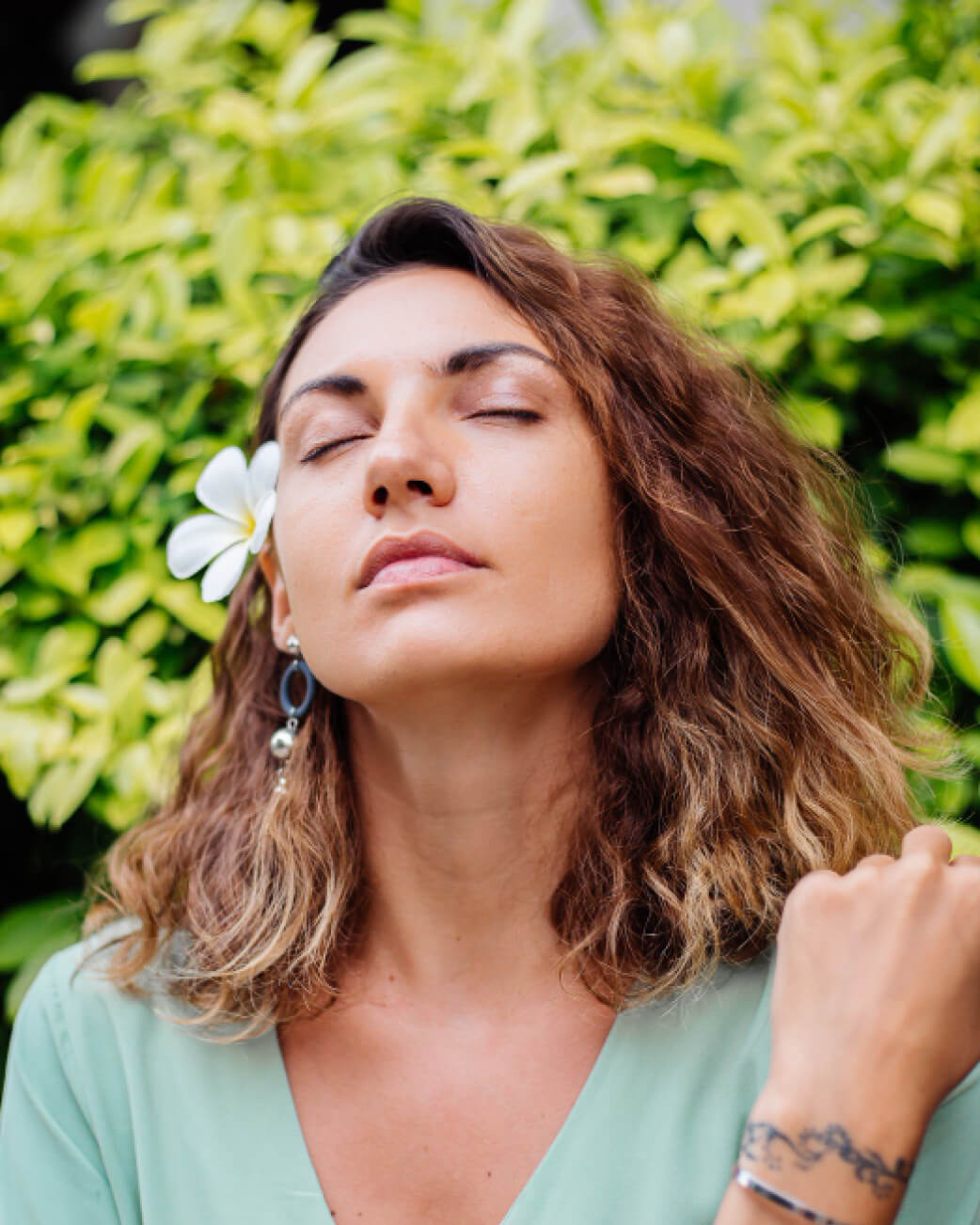 Woman with a flower in her hair against a green leafy background.