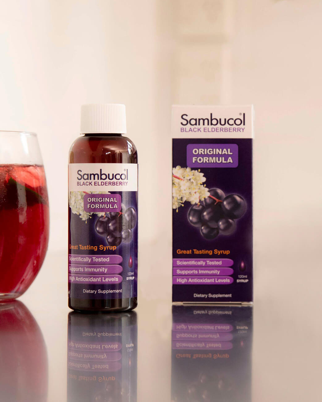 Sambucol black elderberry syrup bottle and packaging on a reflective surface with a glass of red liquid in the background.