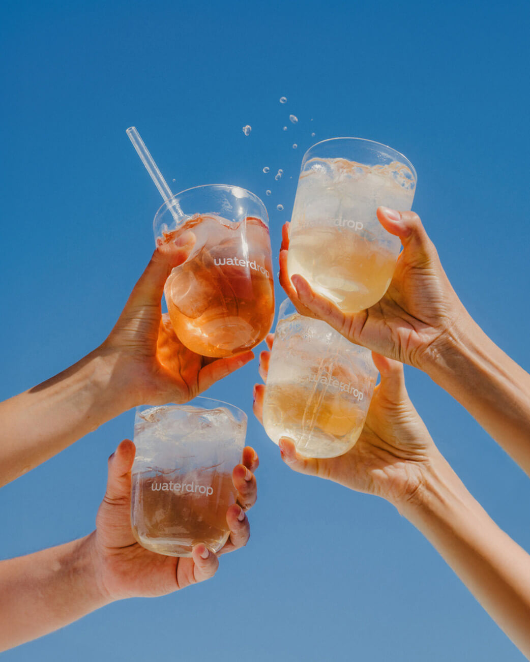 Four hands holding glasses of iced beverages against a clear blue sky.