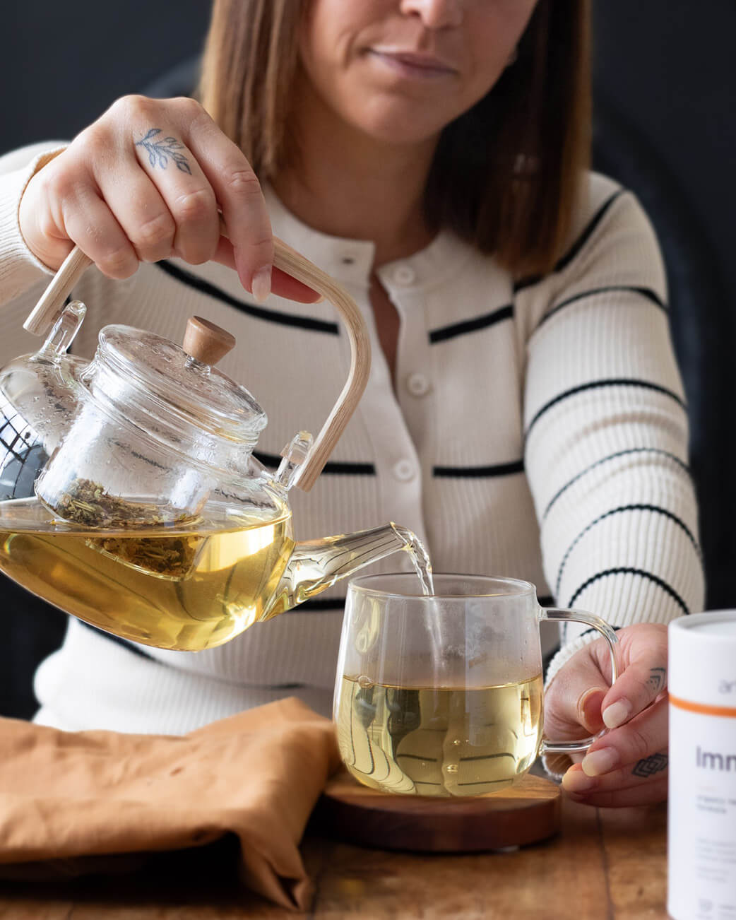 Woman pouring tea from a glass teapot into a glass mug on a wooden table.