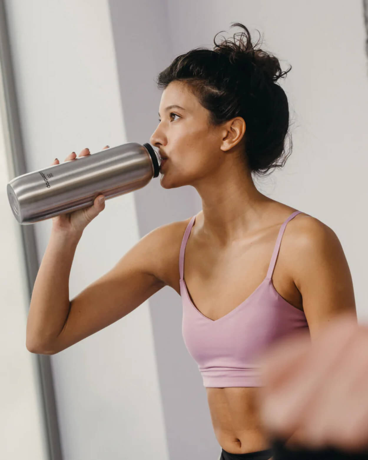 Woman in a pink sports bra drinking from a silver water bottle against a neutral background.