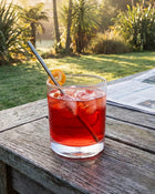 Red cocktail with a lemon wheel and straw in a glass on a wooden table outdoors.