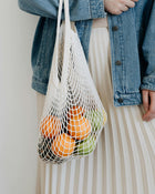 Person holding a string bag with oranges and apples against a neutral background