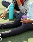 Two water bottles, one blue and one purple, held by people sitting on grass.
