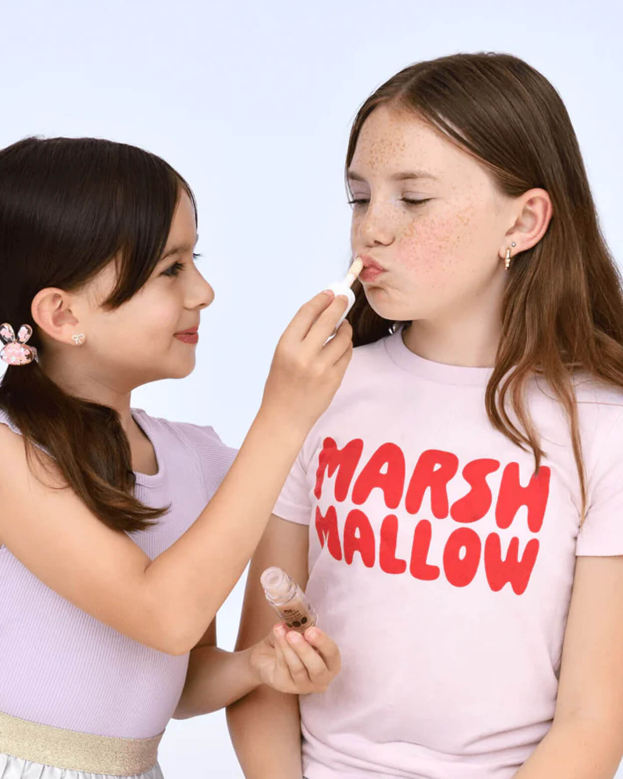 Two young girls with one applying makeup to the other against a light blue background.