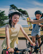 Two children on bicycles with helmets, one being sprayed with sunscreen.