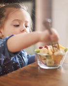 Child reaching for a fruit salad in a bowl at a wooden table.