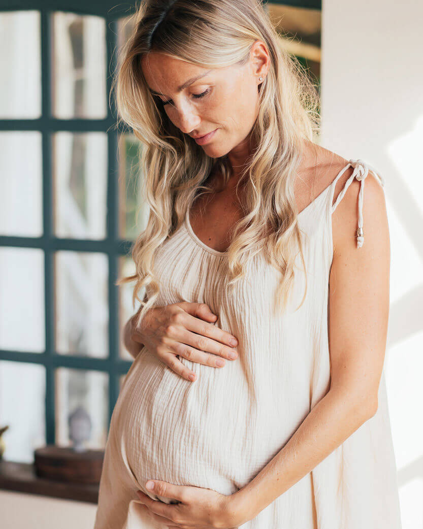 Pregnant woman in a white dress holding her belly indoors.
