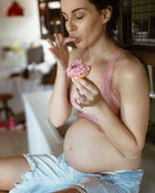 Pregnant woman eating a pink frosted donut in a casual setting.