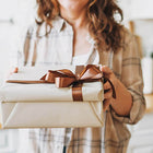 Woman holding out a wrapped gift in neutral coloured paper and brown ribbon.