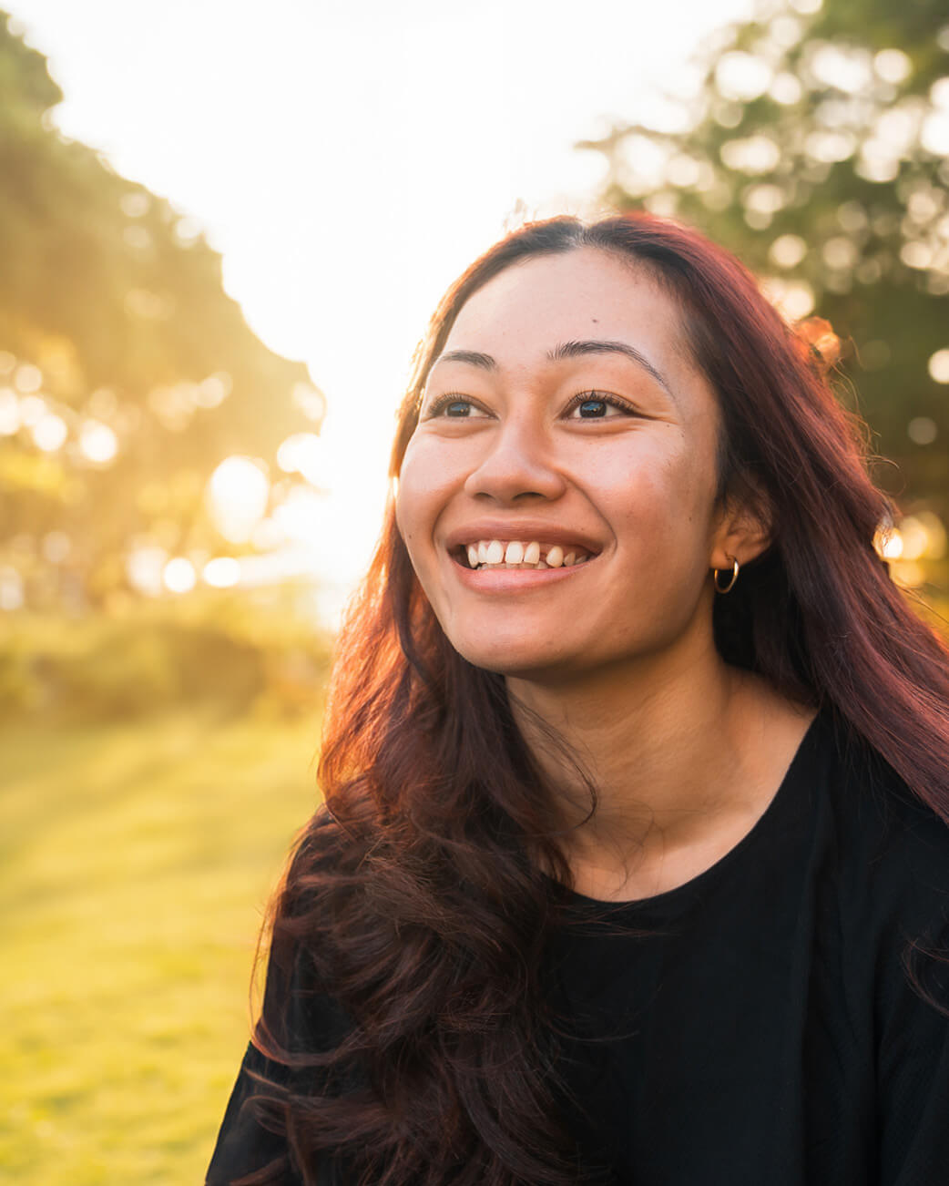 Woman with long dark hair smiling outdoors with a blurred natural background.
