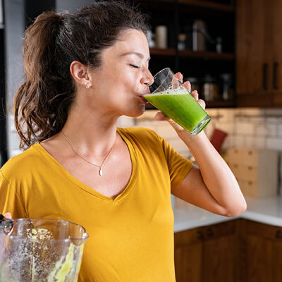 Woman wearing a yellow shirt in a kitchen drinking a green smoothie.