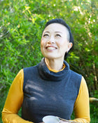 Woman wearing a blue vest over a yellow shirt, standing outdoors with greenery in the background.