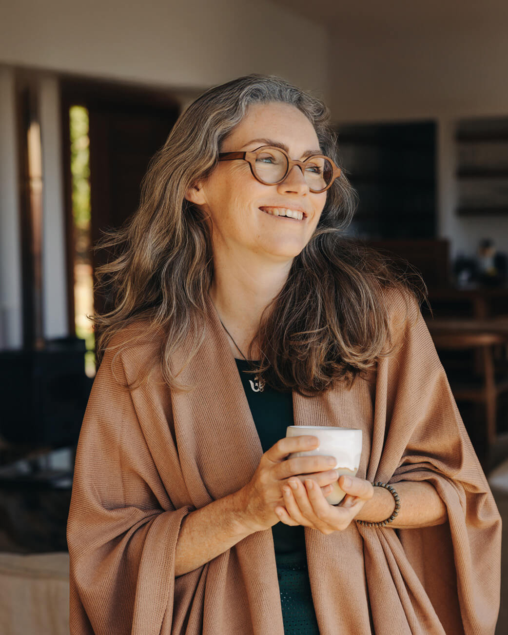 Woman wearing glasses and a brown shawl holding a white mug indoors.