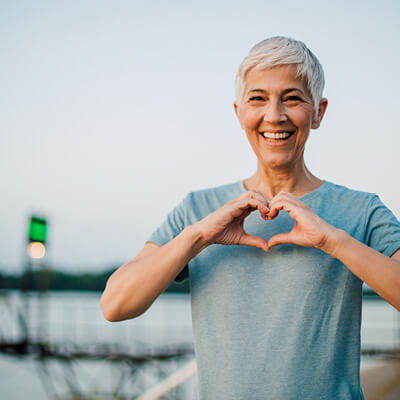 Woman making a heart shape with her hands outdoors by a lake.
