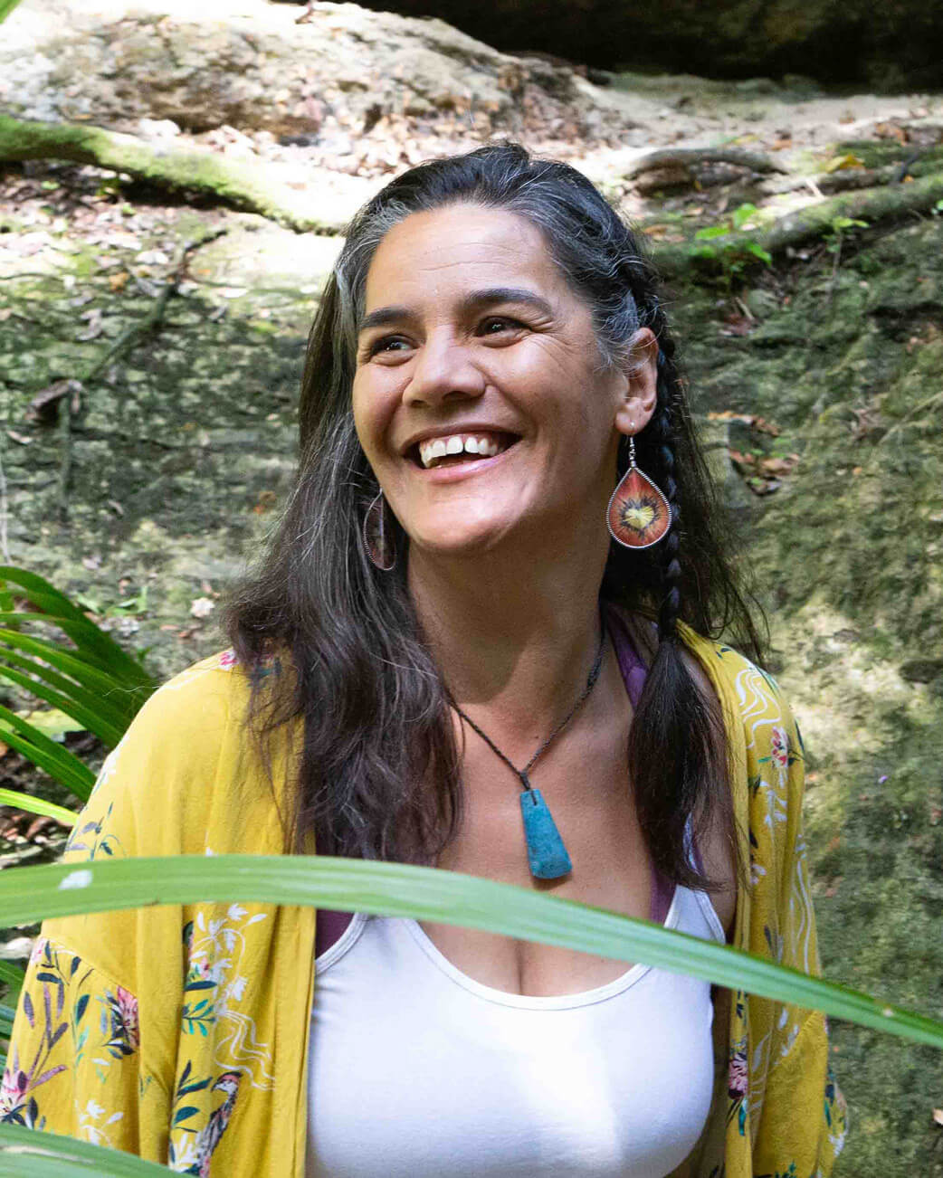 Woman smiling outdoors with greenery and a rock formation in the background.