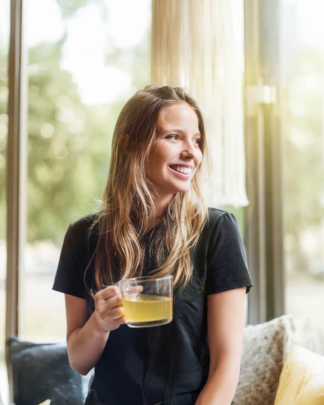 Woman holding a mug of yellow liquid, smiling by a window.