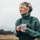 Woman in a green sweater and overalls holding a white mug outdoors.