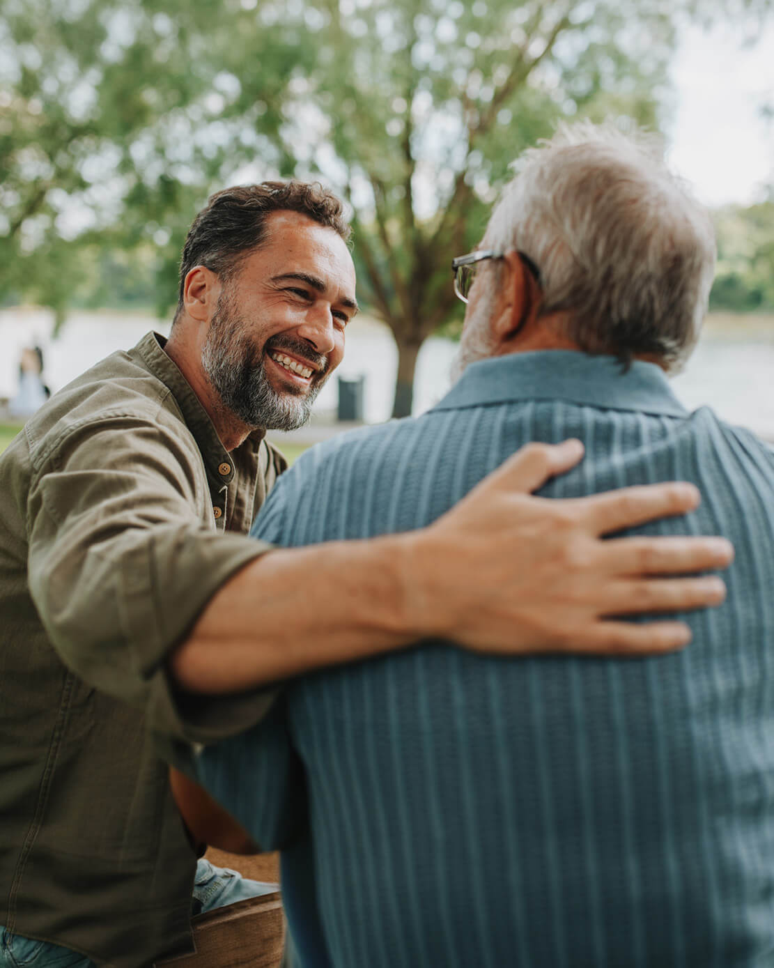 Two men embracing outdoors with trees and a lake in the background.