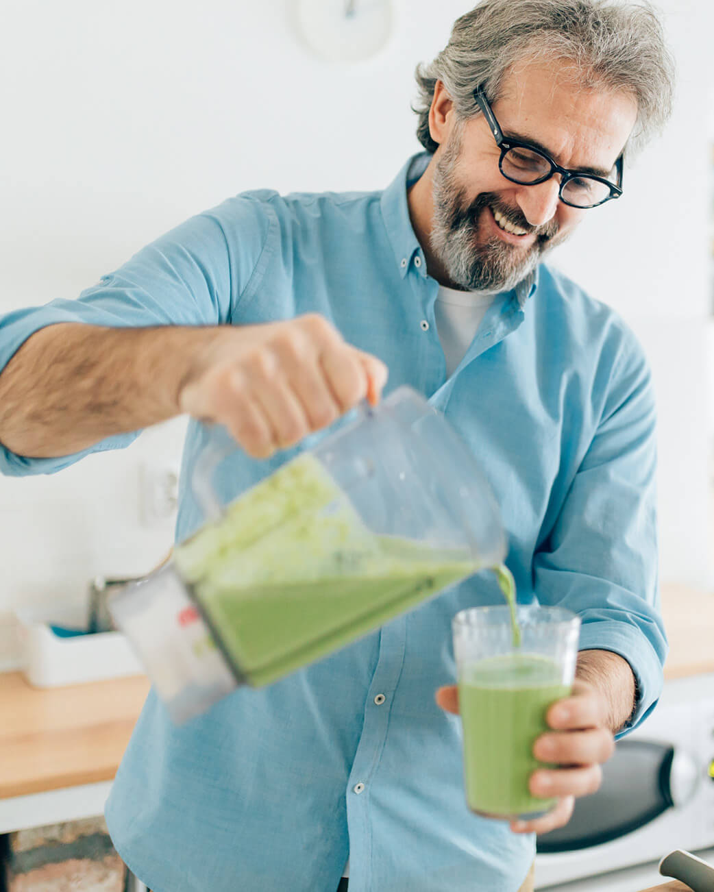 Man pouring green juice from a blender into a glass in a kitchen.
