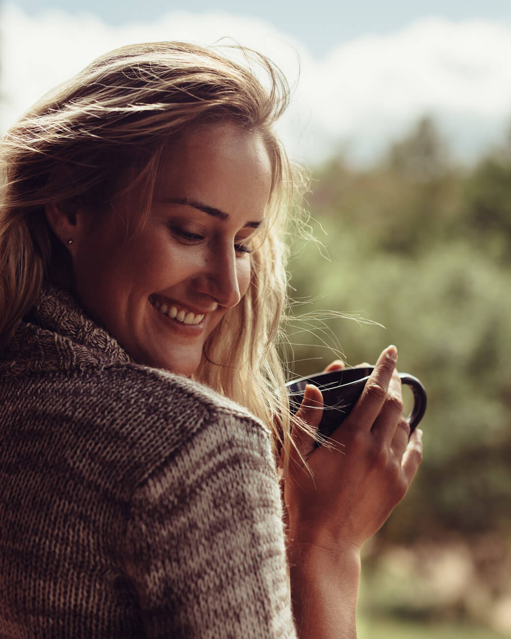 Woman smiling and holding a mug against a blurred natural background.
