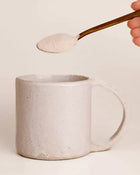 Spoonful of a white powder being added to a ceramic mug on a light background