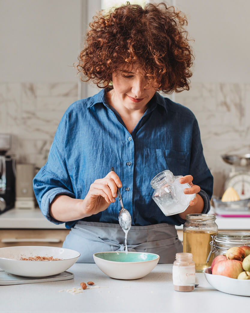 Woman in a kitchen preparing food, holding a jar and spoon.