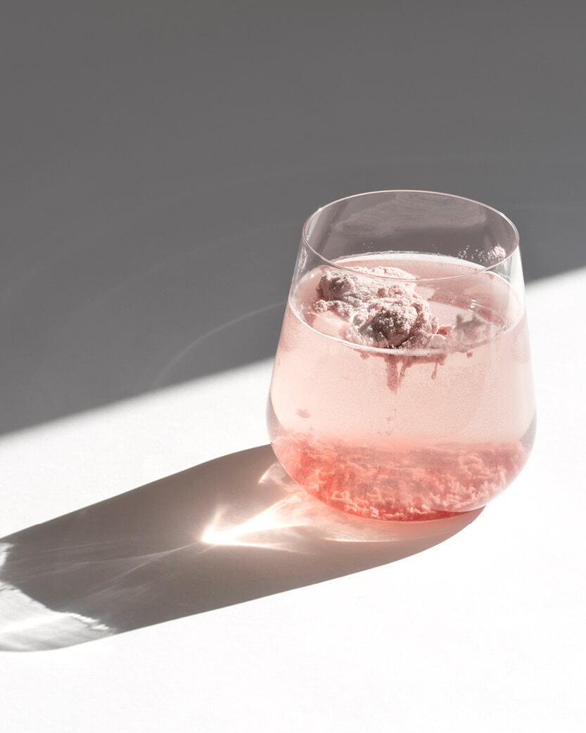 pink collagen protein powder dissolving into a glass of water on a white background with a shadow.