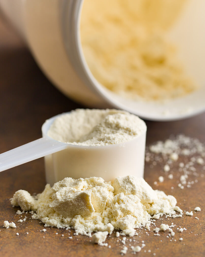 White scoop with Whey protein powder on a wooden surface, with a container of powder in the background.