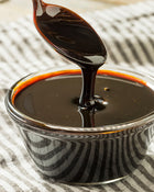Dark syrup being poured from a spoon into a glass bowl on a striped fabric background