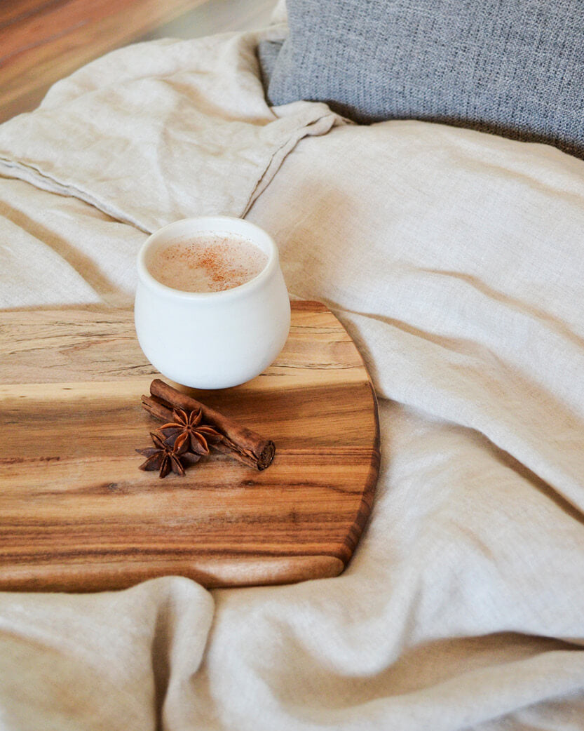 White mug with a warm beverage on a wooden coaster with cinnamon sticks and star anise, placed on a soft beige blanket.