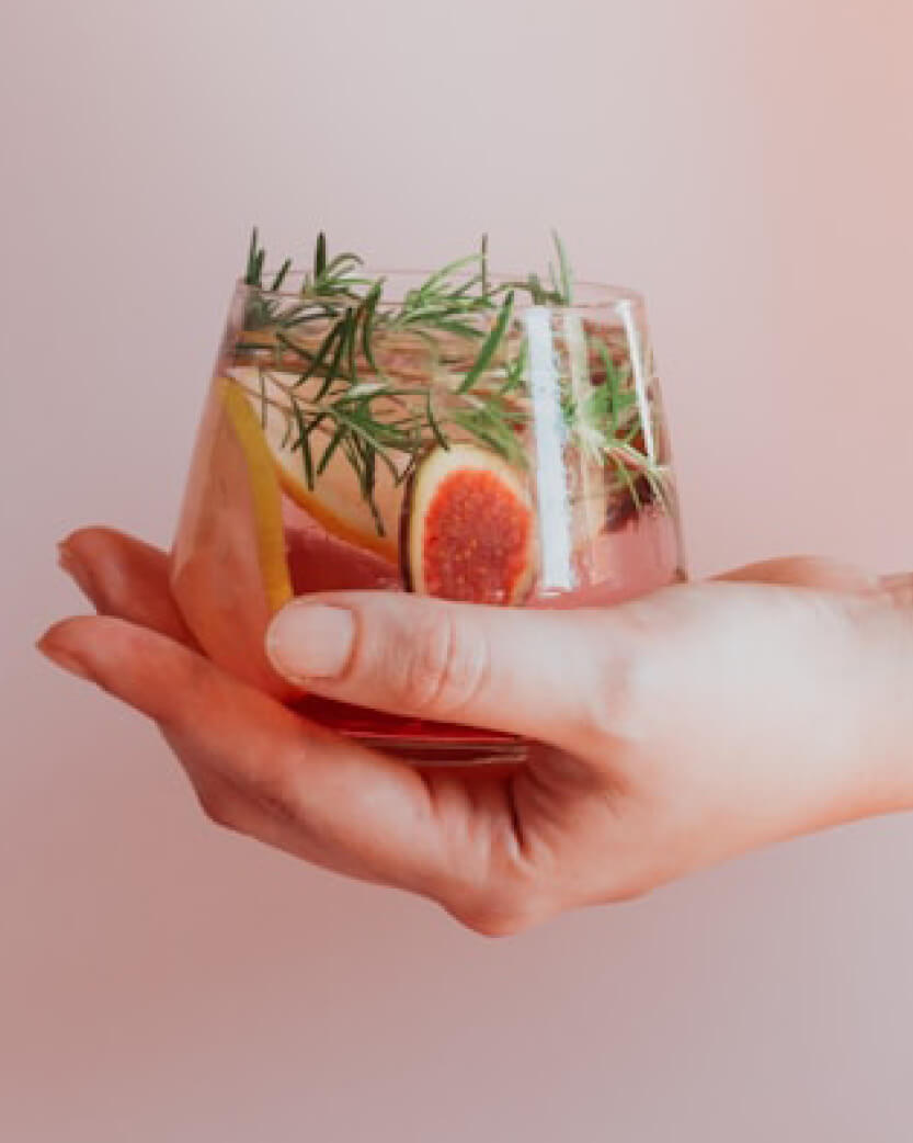 Hand holding a glass of pink cocktail with fruit and herbs against a light pink background