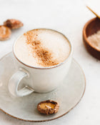 Mushroom latte in a white cup on a saucer with a wooden bowl in the background.
