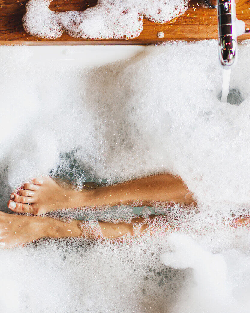 Feet in a bathtub filled with bubbles & water, with a wooden board & faucet visible.