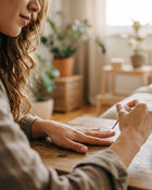 Woman with long brown hair applying nail polish against a blurred background with plants.