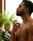 Man spraying cologne on his chest with plants in the background.